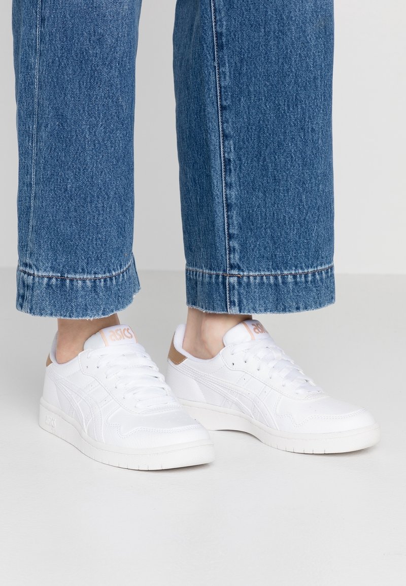 Weiße Sportschuhe mit einer strukturierten Oberseite, die einen hellbraunen Akzent an der Ferse, eine strukturierte Sohle und Schnürsenkel für einen sicheren Sitz aufweisen.