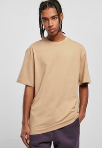 Beige Baumwoll-T-Shirt in Übergröße mit kurzen Ärmeln und Rundhalsausschnitt, das eine glatte Textur aufweist. Kombiniert mit dunkelvioletten Hosen.