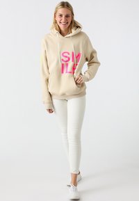 Beige hoodie with bold pink "SMILE" print, drawstrings, and a front pocket. Paired with white skinny jeans and white sneakers.