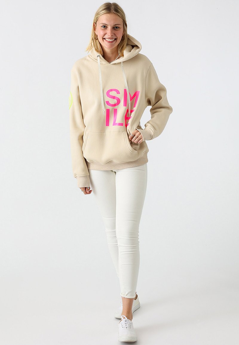 Beige hoodie with bold pink "SMILE" print, drawstrings, and a front pocket. Paired with white skinny jeans and white sneakers.