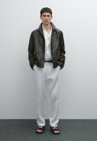 Braune Lederjacke mit Reverskragen, weißes Hemd, hellgraue Leinenhose und schwarze Sandalen. Glatte Texturen und taillierte Passform.