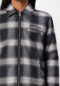 Plaid jacket in black, gray, and white. Features a zip closure, pointed collar, and pocket with metallic hardware. Textured material.