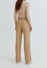 Beige wijd-uitlopende broek met een gladde textuur, hoge taille en riemlussen. Gecombineerd met een fitted lange mouwen top, bekeken vanuit de achterkant.