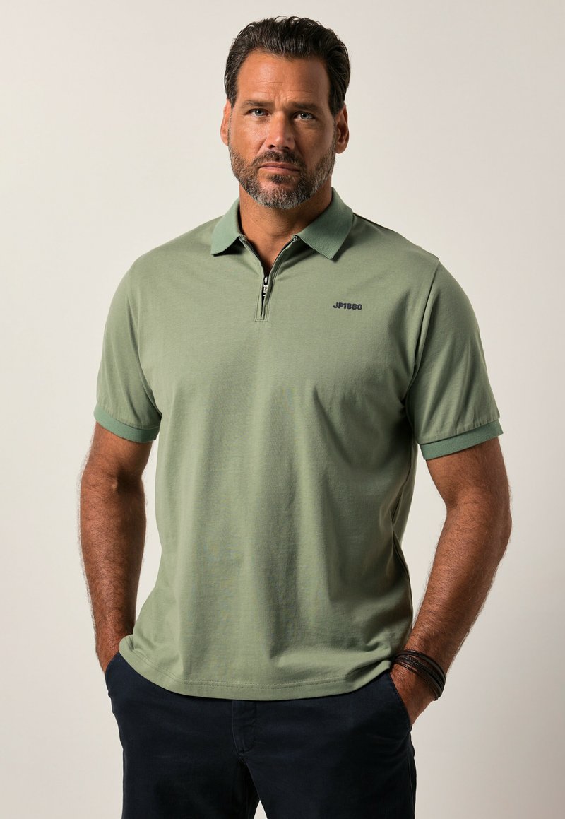 Grön kortärmad pikétröja med dragkedja vid kragen, texturerad bomullstyg och en liten svart logotyp på bröstet.