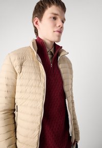 Beige puffer jacket with horizontal quilted pattern, zipper pockets, worn over a textured burgundy sweater with a high collar and knit design.