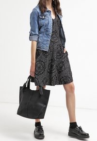 Jeansjacke über einem schwarzen Kleid mit kreisförmigen Mustern, kombiniert mit schwarzen Sneakern und einer großen schwarzen Tragetasche. Strukturierte und lässige Passform.