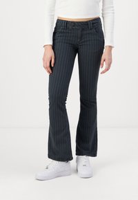 Mujer con pantalones de campana de rayas negras, top corto blanco y zapatillas blancas de pie sobre un fondo blanco.