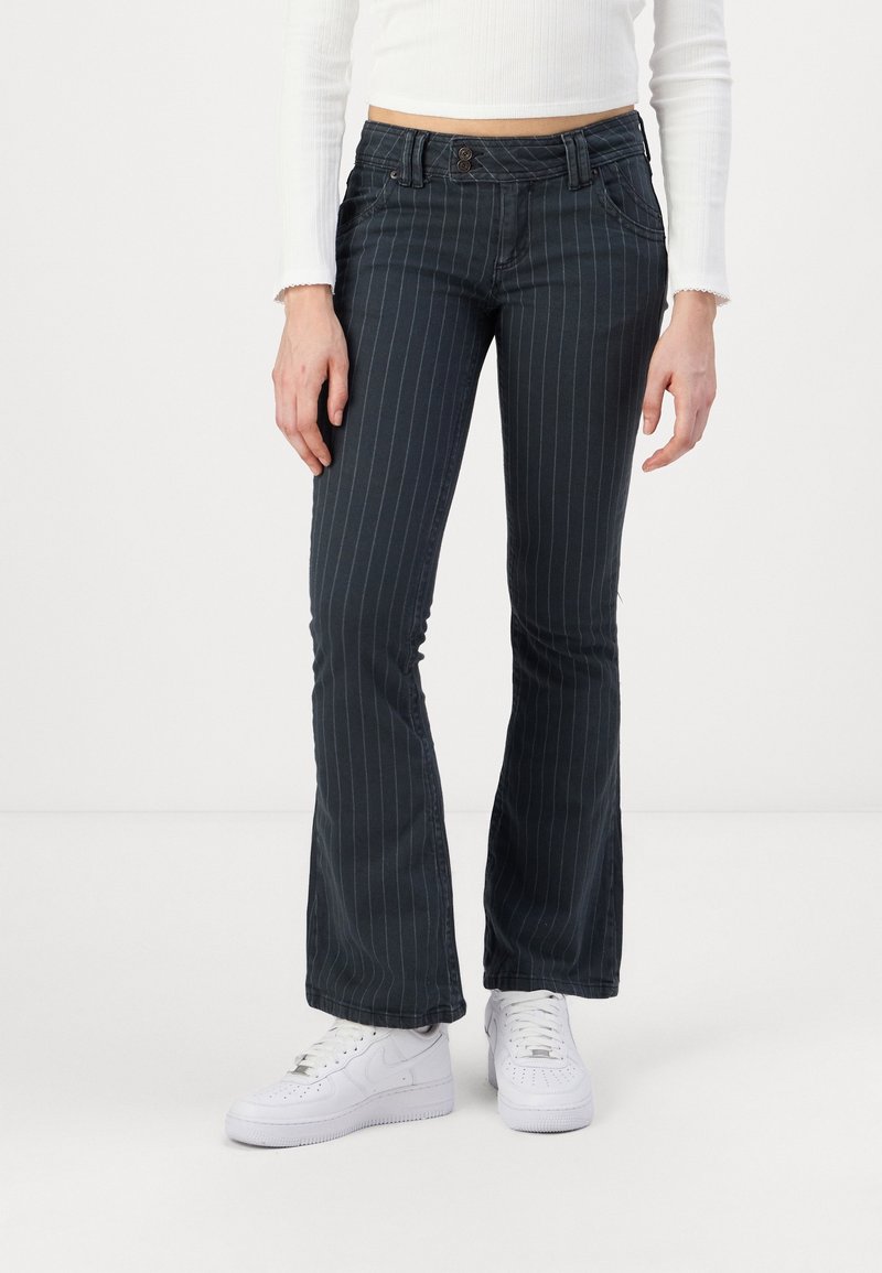 Mujer con pantalones de campana de rayas negras, top corto blanco y zapatillas blancas de pie sobre un fondo blanco.