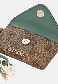 Brązowy portfel o fakturowanej powierzchni z zielonym wnętrzem, złotymi elementami i małym zawieszonym serduszkiem z logo. Zaokrąglone krawędzie z zatrzaskami.