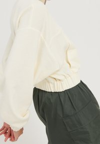 Felpa oversize color crema con vita arricciata, abbinata a pantaloni cargo leggeri verde scuro con tasca laterale.