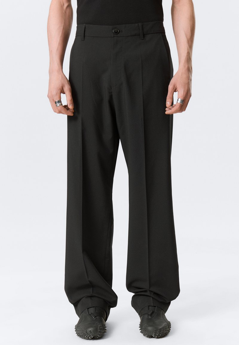 Homme portant un pantalon noir ample, des chaussures noires à semelle cloutée, et des bagues argentées aux deux mains, debout devant un fond clair uni.