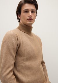 Beige geribbelde coltrui met een aansluitend ontwerp en een gestructureerde brei. De stof lijkt zacht en rekbaar.