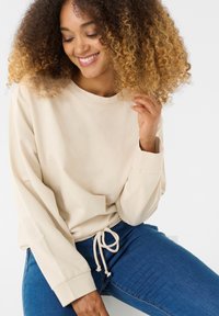 Felpa beige a maniche lunghe con scollatura rotonda e cordoncino all'orlo, abbinata a jeans blu. Il tessuto appare morbido con una texture liscia.