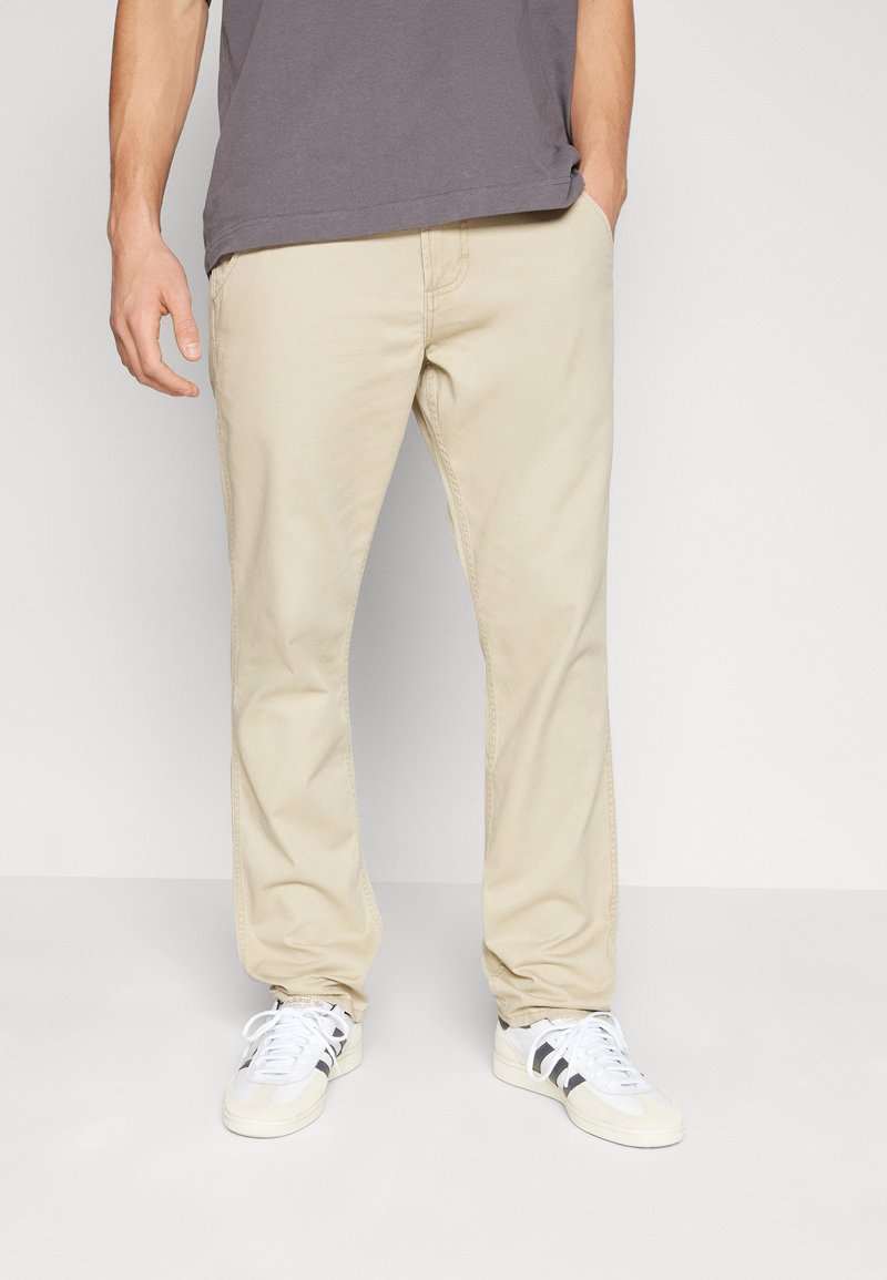 Homme portant un pantalon beige, une chemise grise et des baskets blanches avec des rayures noires, debout devant un fond clair uni.