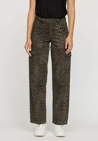 Femme portant un pantalon cargo léopard vert et marron avec de grandes poches et des baskets blanches, se tenant contre un fond uni.