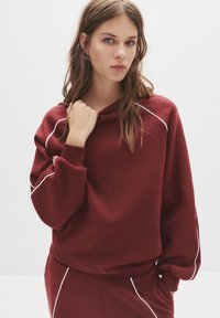 Bordeaux hoodie met een ontspannen pasvorm, witte piping langs de naden, ribgebreide boorden en een trekkoord bij de hals. Zachte stoftextuur.