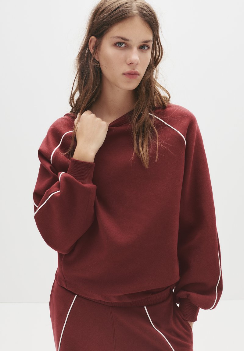 Bordeaux hoodie met een ontspannen pasvorm, witte piping langs de naden, ribgebreide boorden en een trekkoord bij de hals. Zachte stoftextuur.