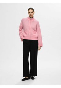 Veste à zip rose avec une finition texturée, dotée de deux poches avant et d'un col montant, associée à un pantalon large noir et des chaussures plates.