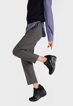 Persona che indossa pantaloni grigio scuro, sneaker platform nere e una camicia a quadri blu con un gilet nero, alzando una gamba su uno sfondo bianco.