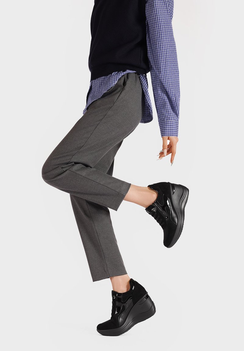 Persona che indossa pantaloni grigio scuro, sneaker platform nere e una camicia a quadri blu con un gilet nero, alzando una gamba su uno sfondo bianco.