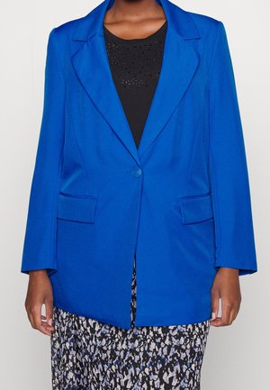 Personne portant un blazer bleu vif à un bouton sur un haut noir avec de petits strass et une jupe à motifs noir, blanc et bleu.