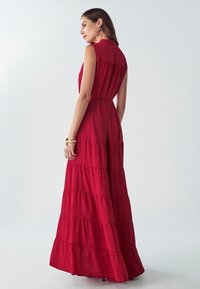 Červené maxi šaty bez rukávov s vrstveným dizajnom a textúrovanou tkaninou. Majú golierový výstrih a zúžený pás, ktorý prechádza do lemu.