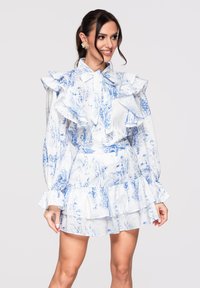 Femme souriante, portant une robe blanche à manches longues avec des volants, ornée de motifs floraux bleus et une jupe à couches, debout devant un fond uni.