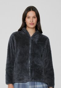 Chaqueta de forro polar con cremallera en gris oscuro, que presenta una superficie suave y texturizada, capucha y mangas largas, combinada con una camisa de cuadros debajo.