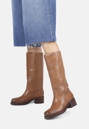 Bottes en cuir marron jusqu'au genou avec un bout arrondi, un petit talon carré, une texture lisse et des détails de couture subtils. Portées avec un denim court.