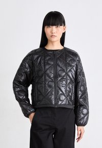 Vrouw met lang steil zwart haar, gekleed in een zwarte gewatteerde jas met cirkel- en lijnpatronen, staand met één hand in haar zak.