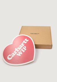 Červená srdcovitá značka s bielym textom "Carhartt WIP", lesklý povrch, umiestnená vedľa hnedého kartónového boxu s logom značky.