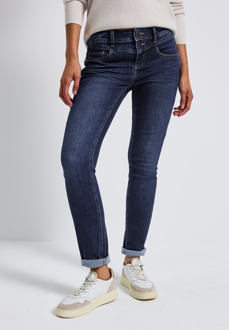 Jean en denim bleu foncé, skinny, avec une taille mi-haute, présentant une fermeture à deux boutons, des coutures discrètes et des ourlets retroussés. Associé à des baskets blanches.