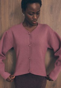 Pulover v barvi praškaste vrtnice z V-izrezom, dolgimi rokavi in petimi gumbi spredaj. Mehka tekstura in nekoliko skrajšan dizajn.