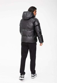 Doudoune noire avec une finition brillante, capuche et design matelassé horizontal. Associée à un pantalon noir et des baskets grises, vue de l'arrière.