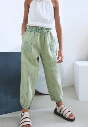 Personne portant un pantalon rayé vert clair avec des poignets élastiques aux chevilles, un haut sans manches blanc à volants et des sandales blanches à semelles noires.