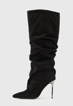 Bota negra de tacón stilettos hasta la rodilla, hecha de tela suave, con diseño arrugado, punta puntiaguda y tacón metálico brillante.