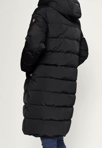 Person med lång svart kviltad vinterjacka med huva, händerna i fickorna, och blå jeans, vänd bort från kameran mot en enfärgad bakgrund.