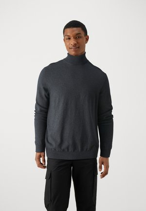 Pull à col roulé gris foncé en tissu tricoté doux, avec des manches longues et un ourlet côtelé. Porté avec un pantalon cargo, fond minimal.