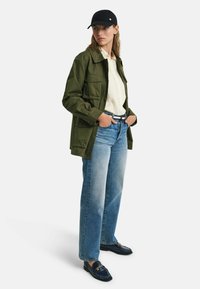 Olivengroene utilityjacket, lichtgestreept shirt, vervaagde blauwe recht-toe-recht-aan jeans en donkere loafers met gouden kettingdetail, zwarte pet.