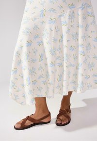 Brune suedine sandaler med krydsende stropper, båret med en hvid nederdel med et blåt blomstermønster og grønne blade.