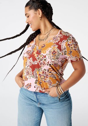Vrouw met lang gevlochten haar, gekleed in een kleurrijke bloemen top en blauwe jeans, glimlachend en poserend met handen in de zakken tegen een witte achtergrond.
