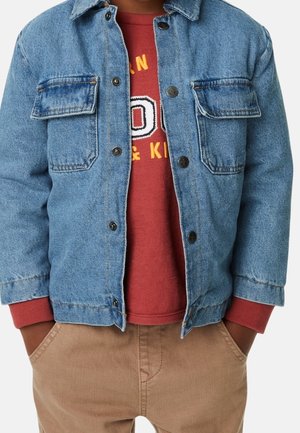 Niño con chaqueta vaquera azul sobre una sudadera roja con texto, manos en los bolsillos de pantalones beige, torso visible, fondo blanco.