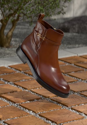 Bruine leren enkelboot met een gladde afwerking, een kleine hak en een gouden accent aan de zijkant. Geplaatst boven een patroon van bestrating.