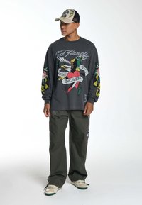Lange mouw grijze shirt met tattoo-stijl graphics; olijfgroene broek; crèmekleurige sneakers met groene zolen; pet met kleurrijke ontwerpen.