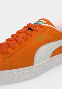 Πορτοκαλί sneaker Puma από σουέτ με λευκή ρίγα, πορτοκαλί κορδόνια και λευκή υφασμένη σόλα σε ουδέτερο φόντο.