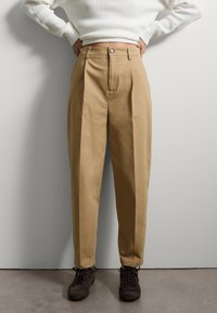 Pantalons kaki à taille haute avec des plis sur le devant, fabriqués à partir d'un tissu robuste. Associés à des chaussures lacées marron foncé et à un pull côtelé blanc.