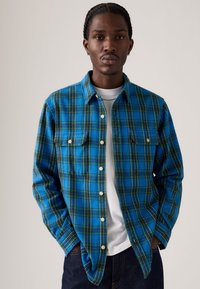 Blauwe en groene geruite flanellen shirt met knoopsluiting aan de voorkant, twee borstzakken en een kraag, gedragen over een wit T-shirt.