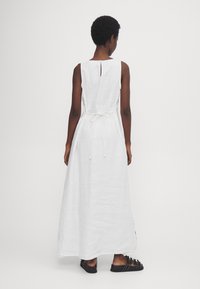 Valkoinen pellava-maksimekko, jossa on pyöreä pääntie, avoin selkä ja säädettävä nauhavyö. Mekossa on virtaava siluetti ja sivuaukot.