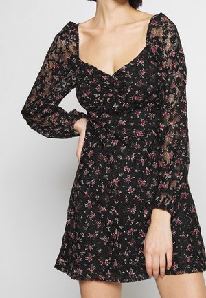 Frau trägt ein schwarzes Spitzenkleid mit Blumenmuster, langen transparenten Ärmeln, gerafftem Oberteil und herzförmigem Ausschnitt, Hand auf der Hüfte.