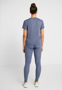 Szary sportowy t-shirt z krótkim rękawem, zestawiony z szarymi legginsami. Wyraźne logo Adidas na plecach, gładka tekstura i dopasowany krój.